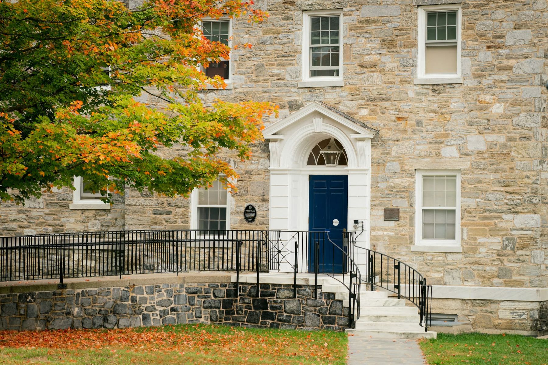 A cut-granite building with a pedimented blue door, black railings and a maple turning orange.