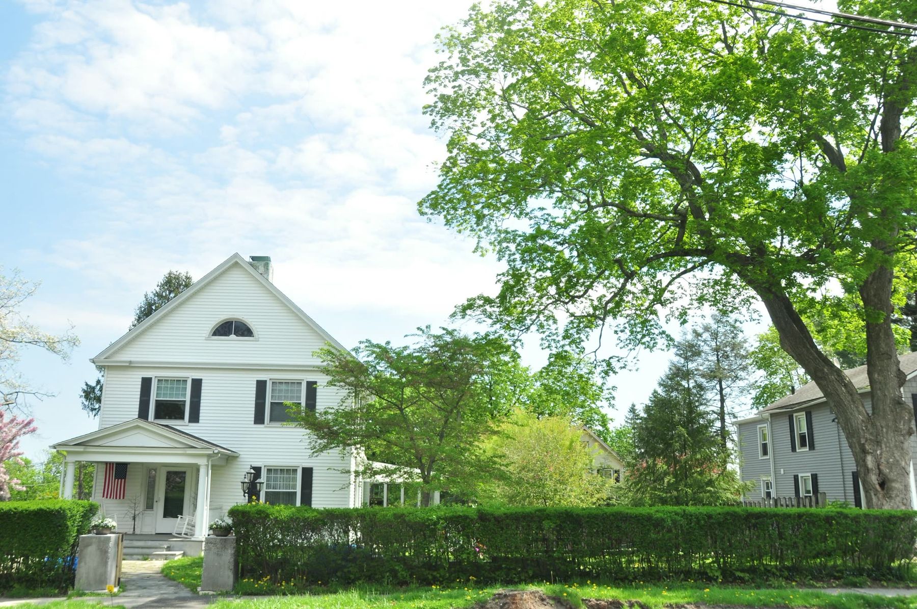 A white two-story colonial with black shutters and an enclosed front porch, behind a clipped hedge under a spring maple.