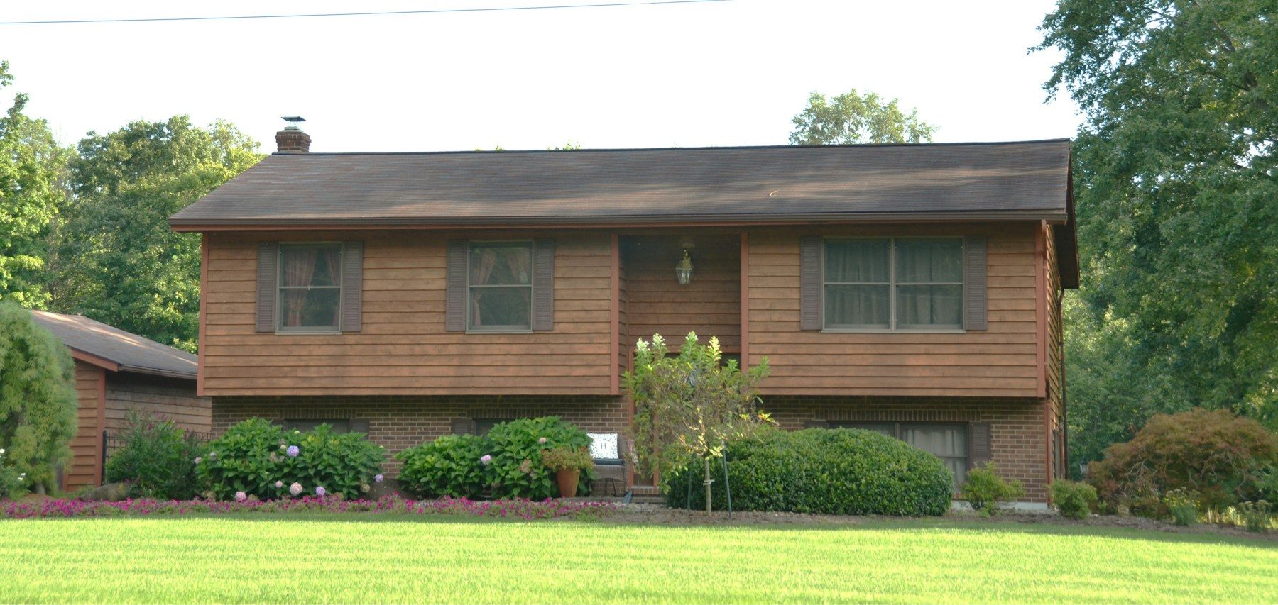 A brown-shingled raised ranch with a brick lower level, shrubs along the front and a wide lawn.