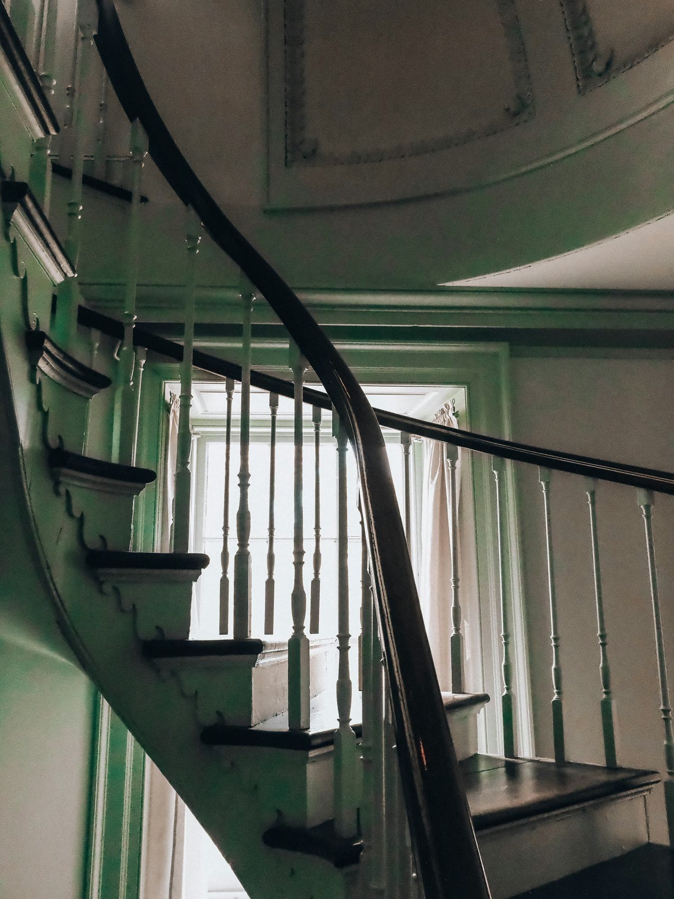 A curved stair with a dark wood rail and white balusters rising past deep green walls toward a tall window.