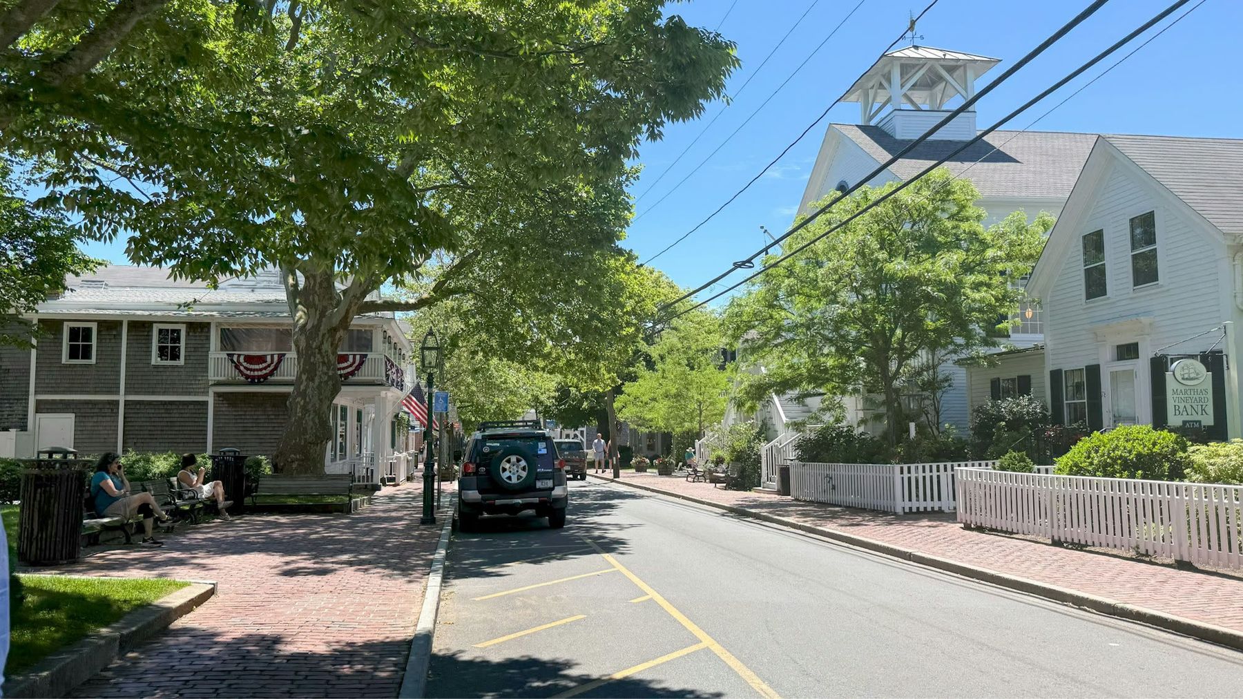 A tree-lined town street with white clapboard houses, picket fences and a church cupola under summer sky.