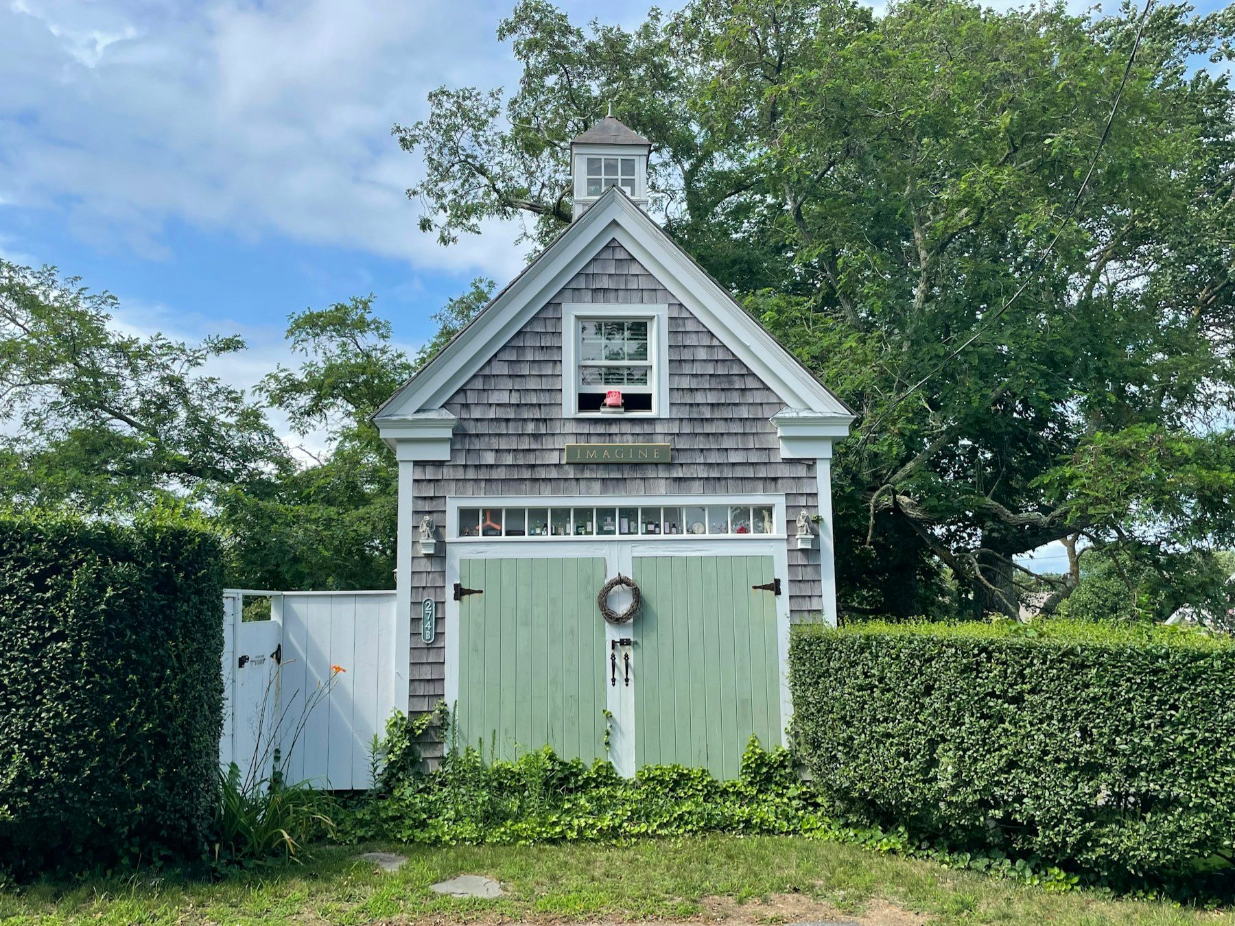 A gray-shingled carriage house with green doors, a cupola and a clipped hedge on either side.