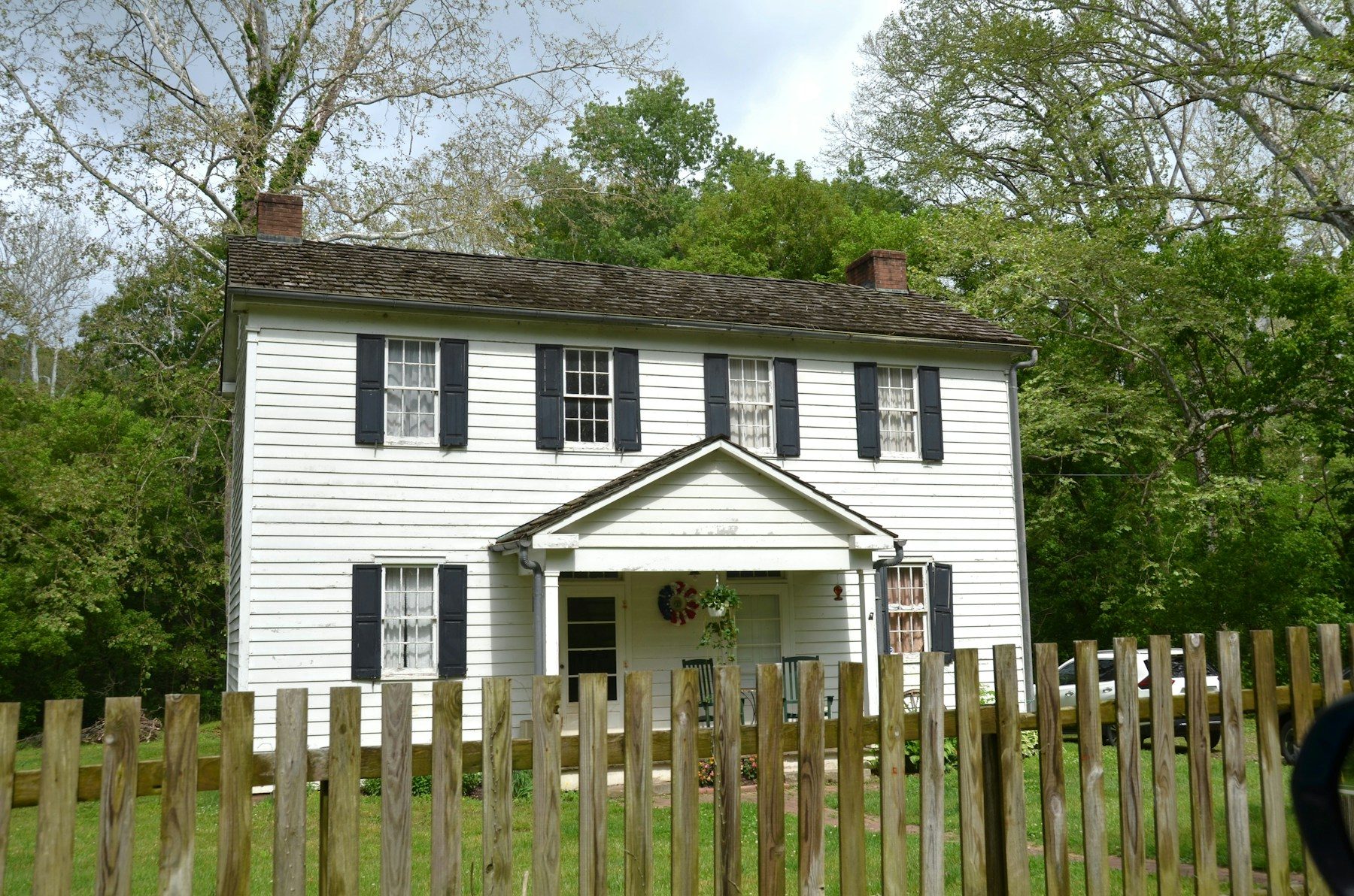 A white side-gabled farmhouse with black shutters, two chimneys and a small center portico behind a split-rail fence.