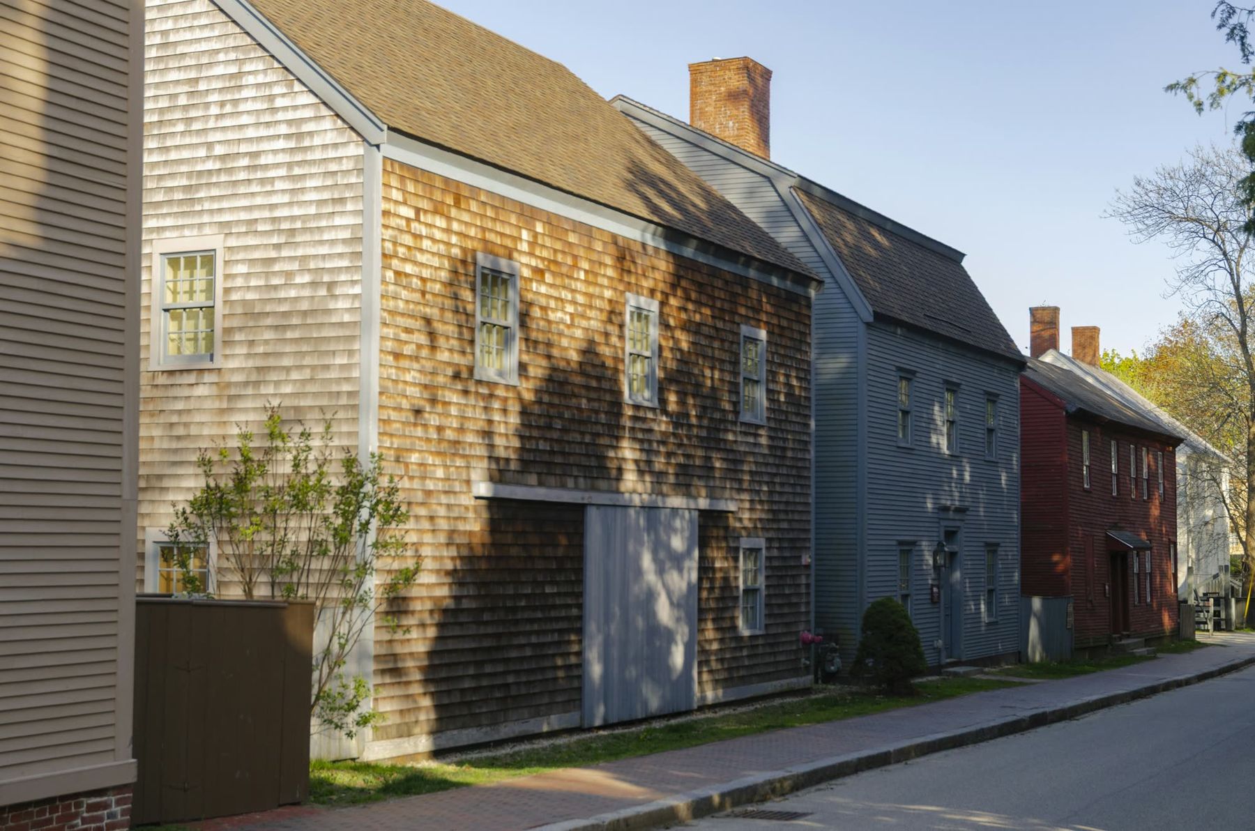 Weathered shingle and clapboard houses in a row along a sunny street, long shadows across the sidewalk.